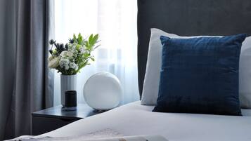A neatly made bed with a blue pillow, a white pillow, and a gray quilt. There is a white vase with flowers on a bedside table.