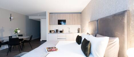 A modern hotel room with a large bed, a dining area, and a kitchenette.