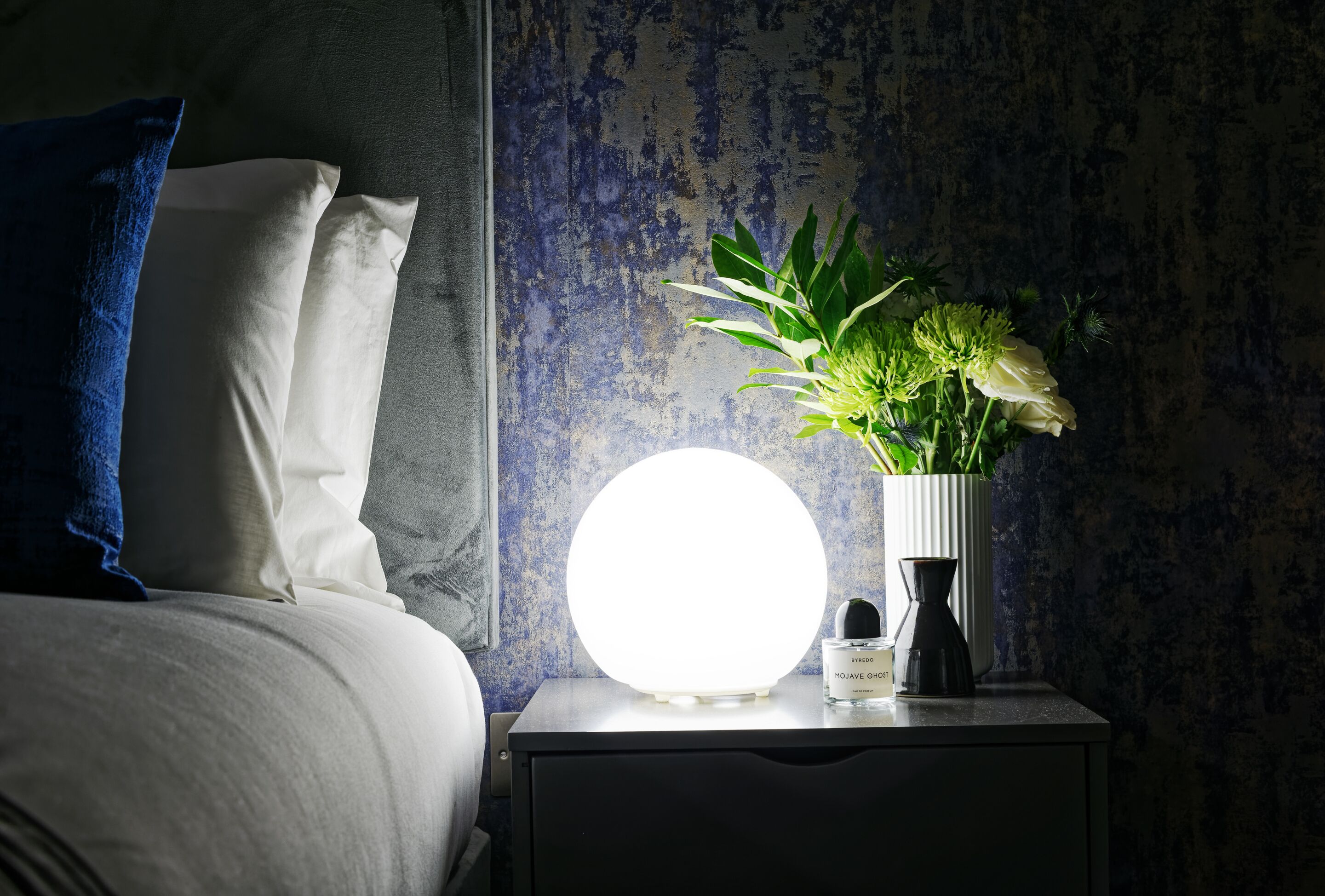 A bedside table with a white vase containing a plant and a bottle of perfume.