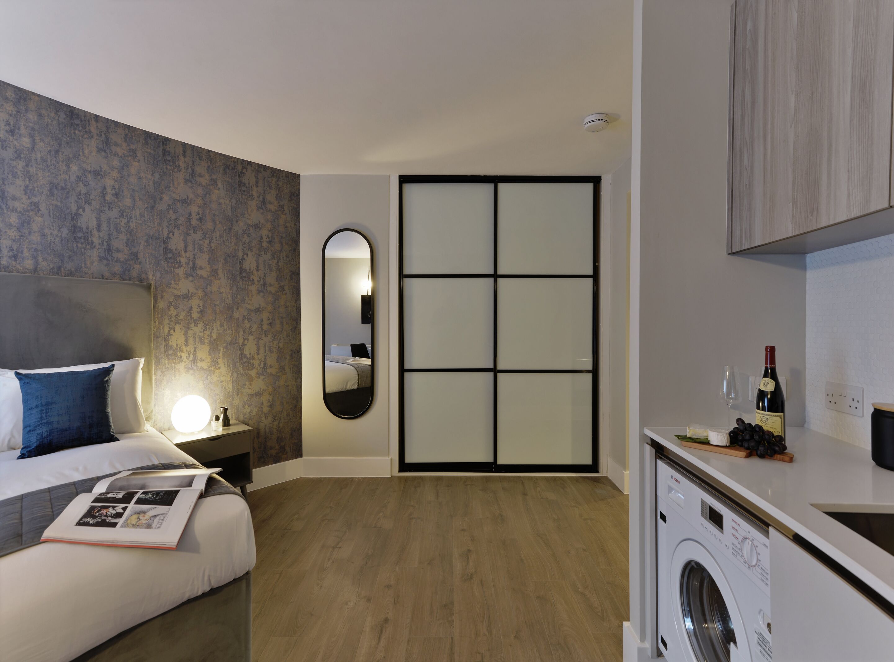 A modern hotel room with a bed, a bedside lamp, a mirror, a wardrobe, a washing machine, and a small kitchen area with a wine bottle and grapes.
