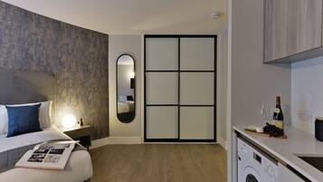 A modern hotel room with a bed, a bedside lamp, a mirror, a wardrobe, a washing machine, and a small kitchen area with a wine bottle and grapes.