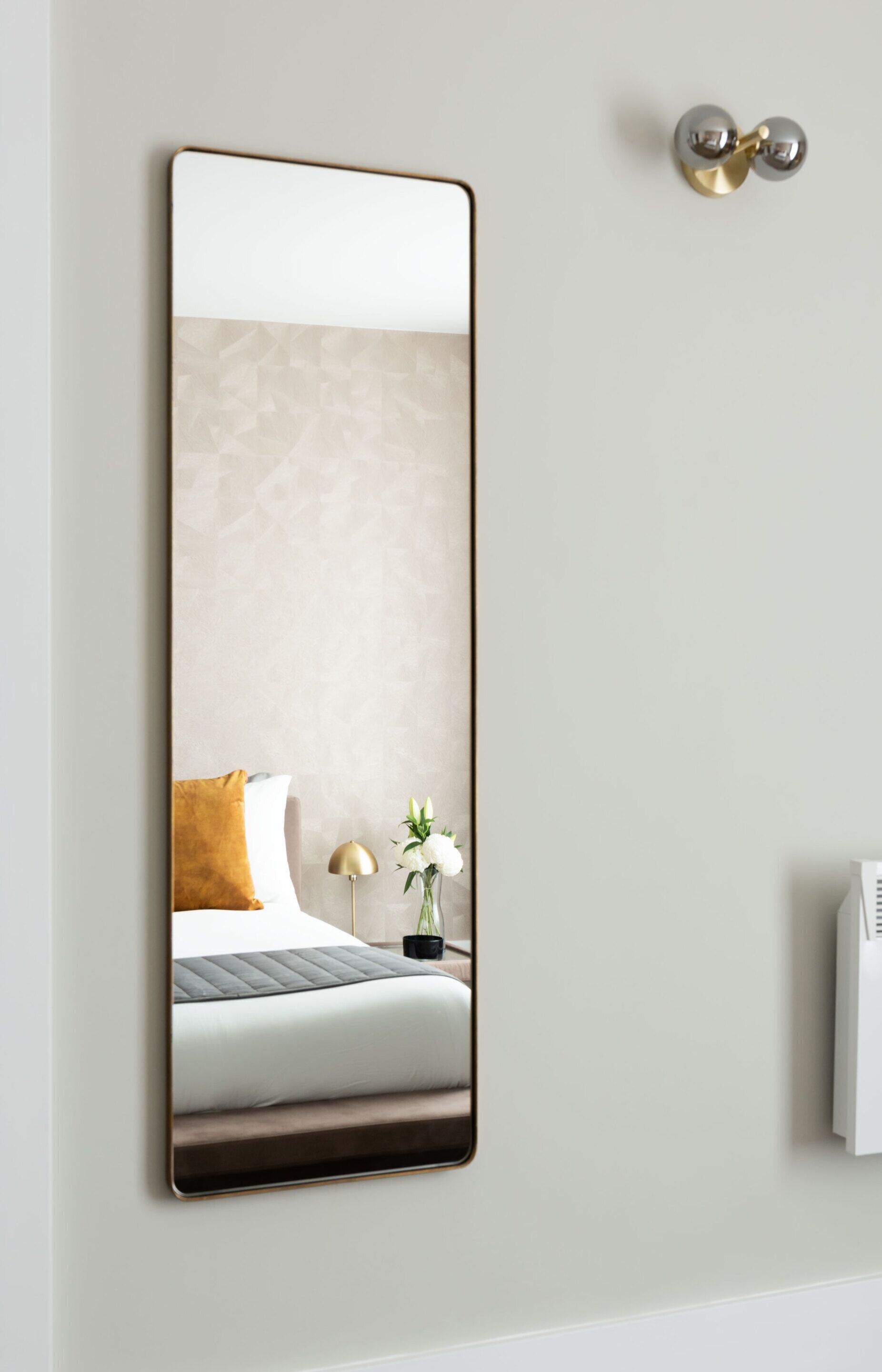 A modern hotel room with a large mirror, a bed with a white and brown duvet, a small bedside table with a lamp, and a wall-mounted air conditioning unit.