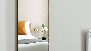 A modern hotel room with a large mirror, a bed with a white and brown duvet, a small bedside table with a lamp, and a wall-mounted air conditioning unit.