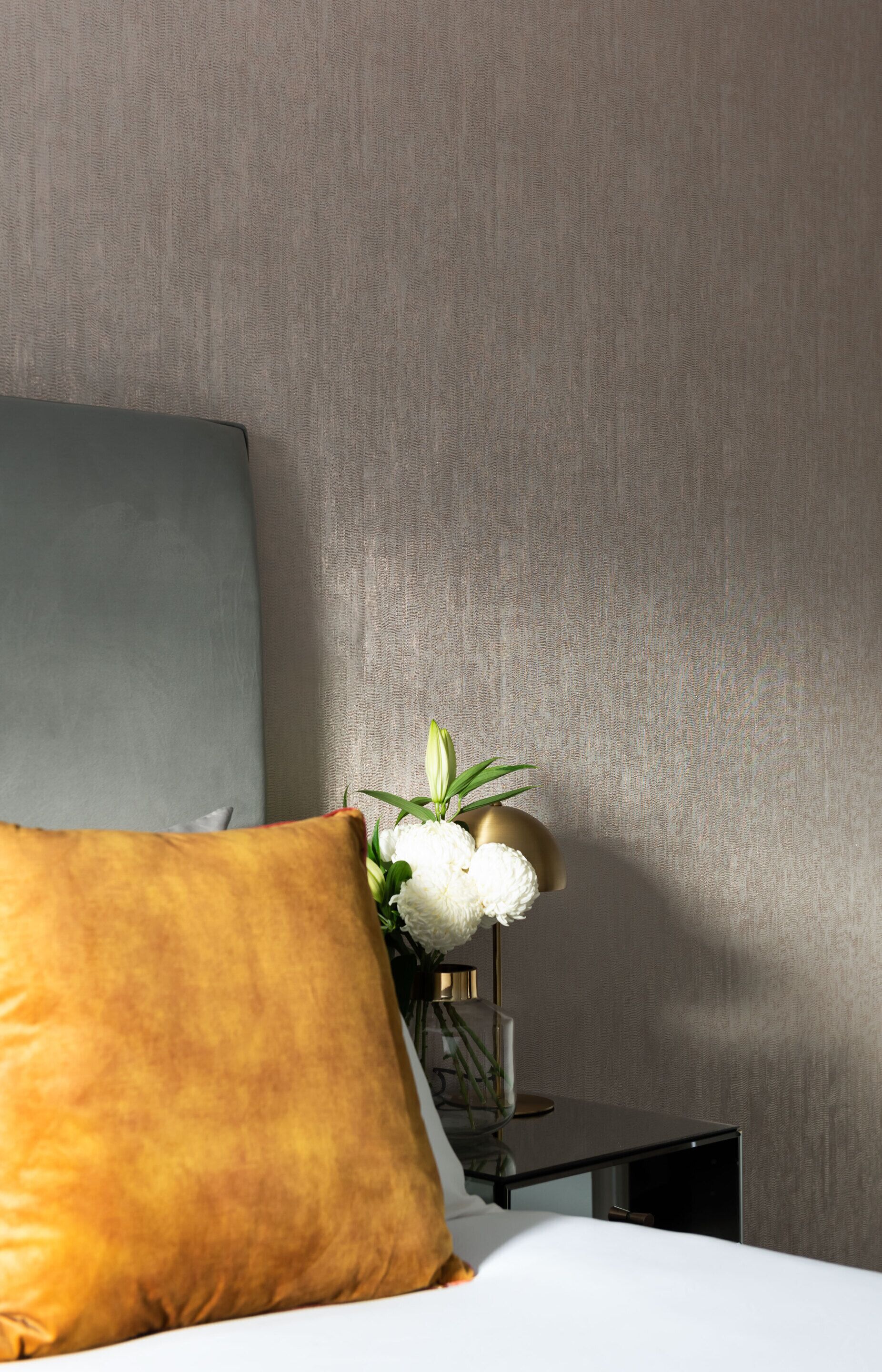 Una cama con cabecera gris, almohada amarilla y una mesita con un florero de flores blancas.