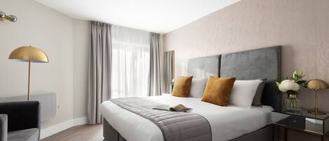 A modern hotel room with a large bed, a bedside table with a lamp, a chair, and a view of the city through large windows.