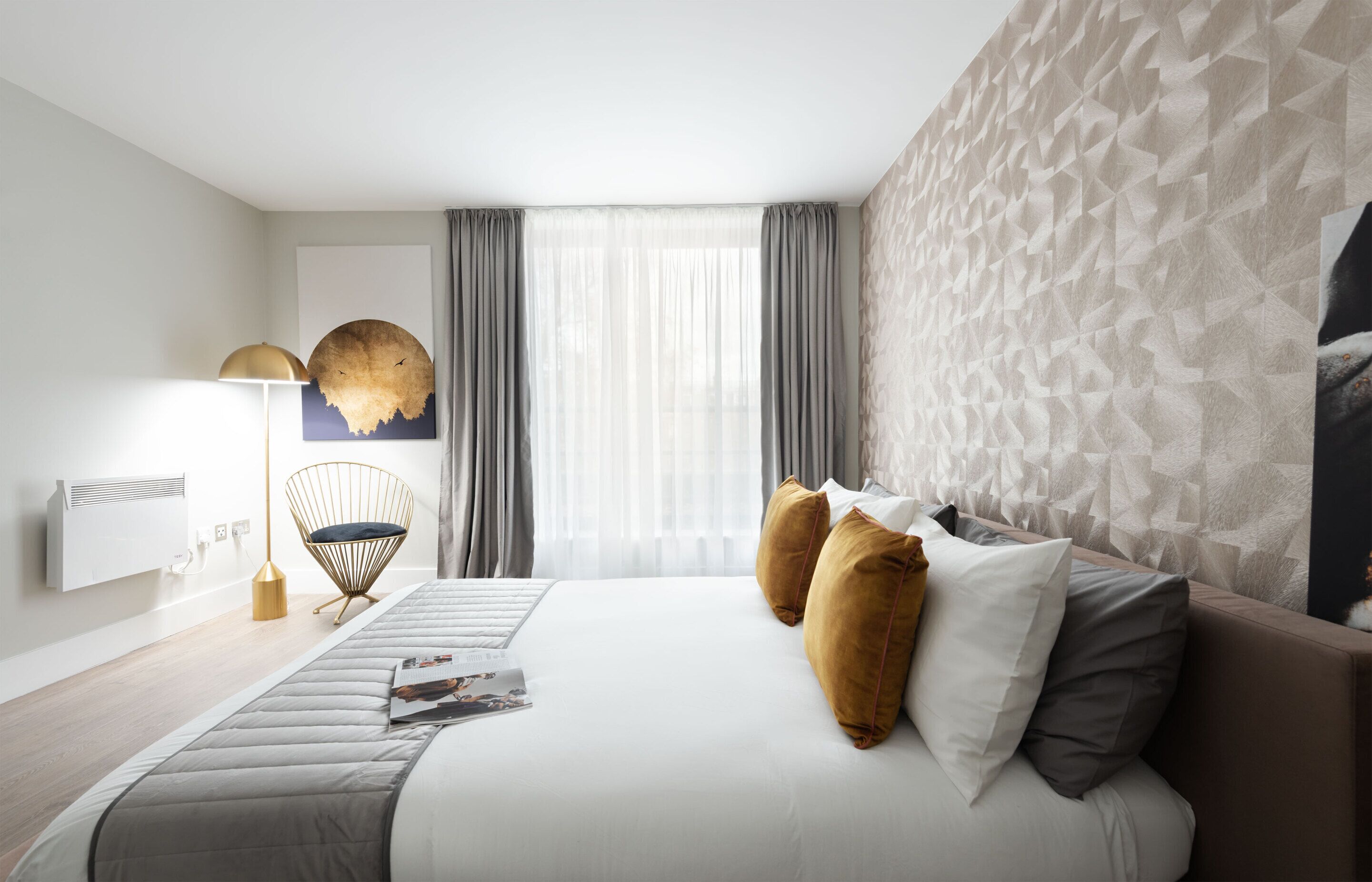 A modern hotel room with a large bed, a round wall clock, a floor lamp, and a view of the city through large windows.
