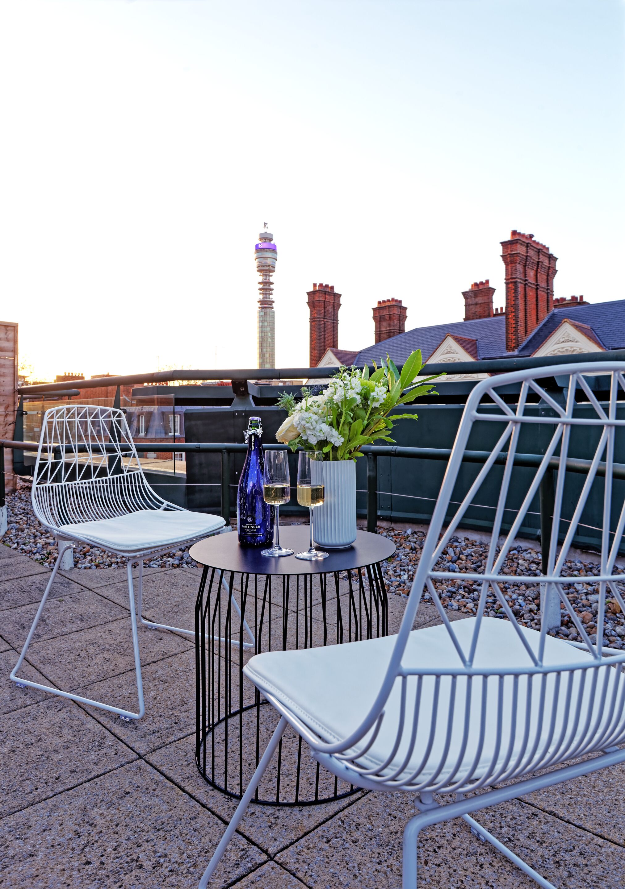 A rooftop terrace with two white metal chairs, a small round table with a bottle and flowers, and a view of a city skyline including a tower and red-roofed buildings.