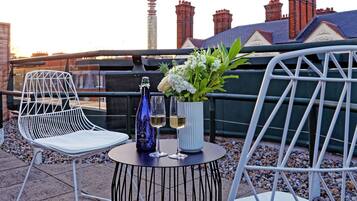 A rooftop terrace with two white metal chairs, a small round table with a bottle and flowers, and a view of a city skyline including a tower and red-roofed buildings.