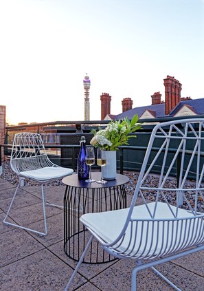 En takterrass med två vita metallstolar, ett litet runt bord med en flaska och blommor, och utsikt över en stadsbild med bland annat ett torn och byggnader med röda tak.