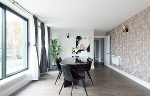 Ett modernt matområde med ett runt bord, svarta stolar, en vas med blommor och ett stort fönster med gardiner.