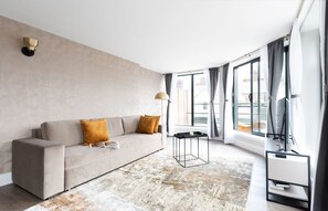 A modern hotel room with a large window, a sofa, a coffee table, and a TV stand. - Three-Bedroom Penthouse (London)