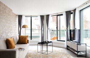 A modern living room with a sofa, a small black side table, a floor lamp, and a TV on a stand. Large windows with curtains provide a view of the outside. - Three-Bedroom Penthouse (London)