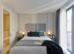 Une chambre d’hôtel avec un grand lit, deux tables de chevet avec des lampes, une fenêtre avec des rideaux et une vue sur les bâtiments environnants.