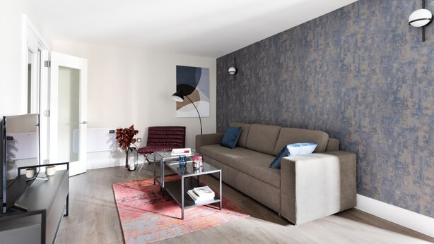 A modern living room with a sofa, coffee table, and a red chair. There is a glass table with a vase and a lamp. The room has a patterned wall and a door leading to another room. - Two-Bedroom Apartment near Oxford Street (London)