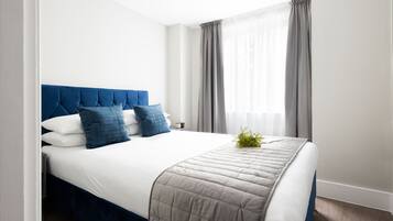 A neatly made bed with a blue headboard, white bedding, and a grey quilt.