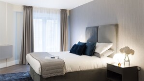 A modern hotel room with a large bed, a bedside table with a lamp, and a view of the outdoors through sheer curtains. - Two-Bedroom Apartment near Oxford Street (London)