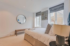 Een slaapkamer met een bed, een bankje, een ronde spiegel en een raam met gordijnen.