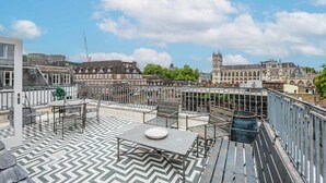 Een terras op het dak met een visgraatvloer, buitenmeubilair en uitzicht op historische gebouwen, waaronder een toren.