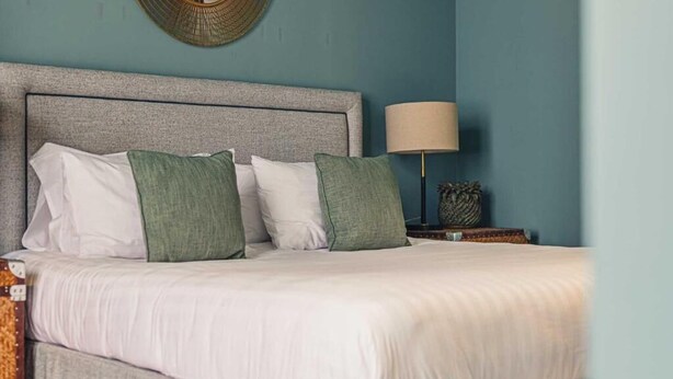 Een net opgemaakt bed met witte en groene kussens, een grijze hoofdbord en een bedlampje.