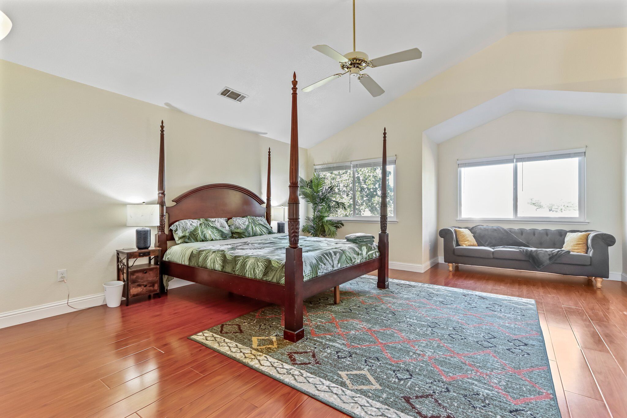 A spacious bedroom with a four-poster bed, a ceiling fan, a large window, and a gray sofa.