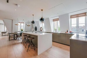 A modern kitchen with a central island, bar stools, pendant lighting, and large windows. - Three-Bedroom Duplex Penthouse (London)