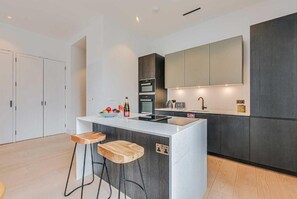 Une cuisine moderne dotée d’un îlot central, de tabourets de bar, d’un four encastré et d’un grand miroir au-dessus du plan de travail.