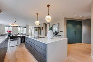 A modern kitchen with a central island, pendant lighting, and a view of the dining area. - Four Bedroom Duplex Penthouse (London)