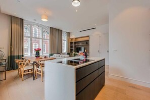 A modern kitchen with a central island, dining area with wooden chairs, and a large window with curtains. - Four Bedroom Duplex Penthouse (London)
