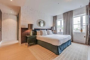 A hotel room with a large bed, a nightstand, a mirror, and a window with curtains. - Four Bedroom Duplex Penthouse (London)
