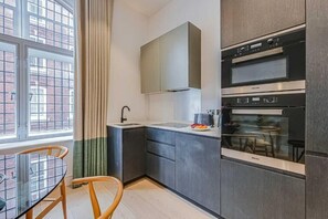 A modern kitchen with a built-in oven, a window with curtains, and a dining area with a glass table and wooden chairs. - Four Bedroom Duplex Penthouse (London)
