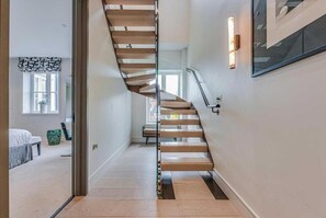 Un escalier moderne avec des marches en bois et une rampe en verre, menant à une chambre à coucher équipée d’un lit et d’une fenêtre.