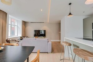 A modern living room with a sofa, a dining table, and a TV. There is a kitchen area with a counter and stools. - Two-Bedroom Superior Apartment (London)