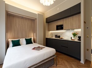 A modern bedroom with a large bed, a nightstand, a built-in desk, and a kitchenette with a microwave. - Standard Studios in South Kensington (London)