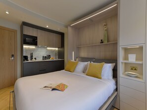 Une chambre d’hôtel moderne équipée d’un lit, d’une petite kitchenette et d’une étagère intégrée sur laquelle est posé un vase.