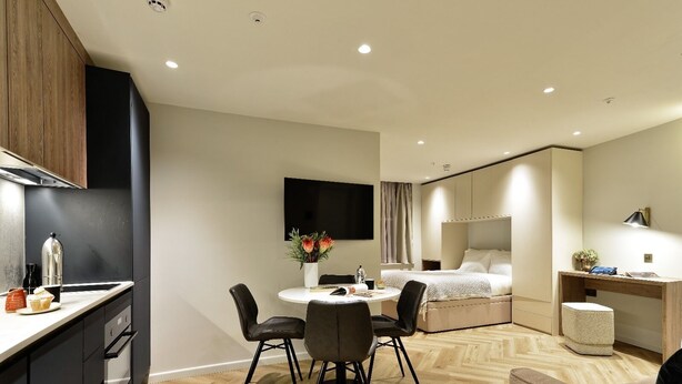Una habitación de hotel moderna con kitchenette, comedor, cama y escritorio con lámpara.