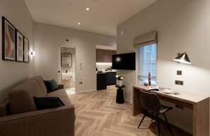 A modern living room with a sofa, a desk, and a TV. There is a kitchen in the background with a window and a TV. - Duplex One Bedroom Apartment (London)