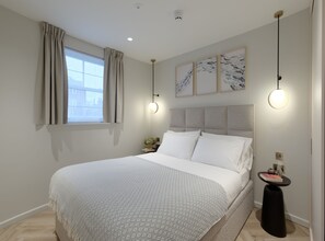 A modern bedroom with a large bed, a window with curtains, a bedside table with a vase, and wall-mounted artwork. - Duplex One Bedroom Apartment (London)