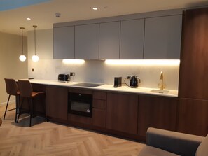 A modern kitchen with a built-in oven, a coffee maker, and a sink. There are two bar stools and pendant lights above the counter. - Duplex One Bedroom Apartment (London)