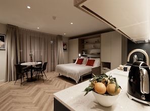 Ein modernes Hotelzimmer mit Essbereich, einem Bett mit roten Kissen, einer Kaffeemaschine und einer Fruchtschale auf der Ablagefläche.