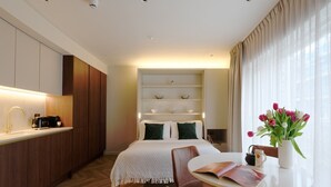 Een moderne hotelkamer met een bed, nachtkastjes, een zithoek met een tafel en stoelen, een kitchenette met een spoelbak en apparatuur, en een groot raam met gordijnen.