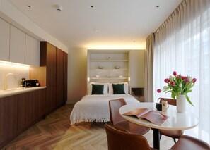 Ein modernes Hotelzimmer mit Bett, Nachttischen, einer Essecke mit Tisch und Stühlen, einer Küchenzeile mit Spüle und Geräten sowie einem großen Fenster mit Vorhängen.