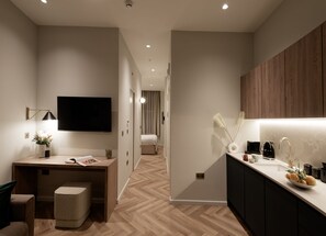A modern hotel room with a wooden floor, a desk with a lamp, a flat-screen TV, a bed, and a kitchenette with a coffee machine. - Superior Studio in Notting Hill (London)