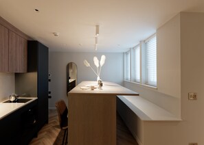 A modern kitchen with a central island, wooden cabinets, a built-in bench, and a large window with blinds. - Superior Studio in Notting Hill (London)