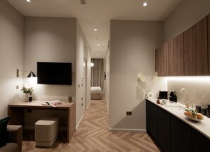 Une chambre d’hôtel moderne avec un plancher en bois, un bureau avec une lampe, un téléviseur à écran plat, un lit et une kitchenette équipée d’une machine à café.