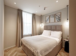 A modern bedroom with a large bed, a nightstand, a wall-mounted lamp, and a window with curtains. - Superior Studio in Notting Hill (London)