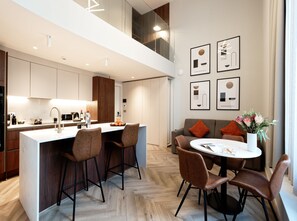 A modern kitchen with a central island, bar stools, and a dining table with chairs. There are framed artworks on the wall and a vase with flowers on the dining table. - Superior Studio in Notting Hill (London)