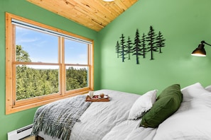 Quarto com uma janela grande, uma cama com roupa de cama branca, um travesseiro verde e uma luminária na parede.