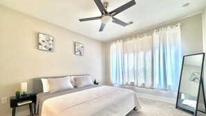 Ein Schlafzimmer mit Bett, Deckenventilator, Nachttisch, Fenster mit Vorhängen und Spiegel.