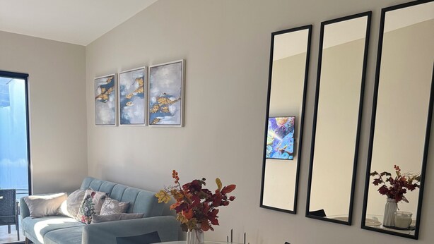 A modern living room with a glass dining table, black chairs, and a grey sofa. There are framed artworks on the wall and a vase with flowers on the table.