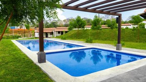 A rectangular swimming pool with a tiled edge, surrounded by a grassy area and a pergola structure.